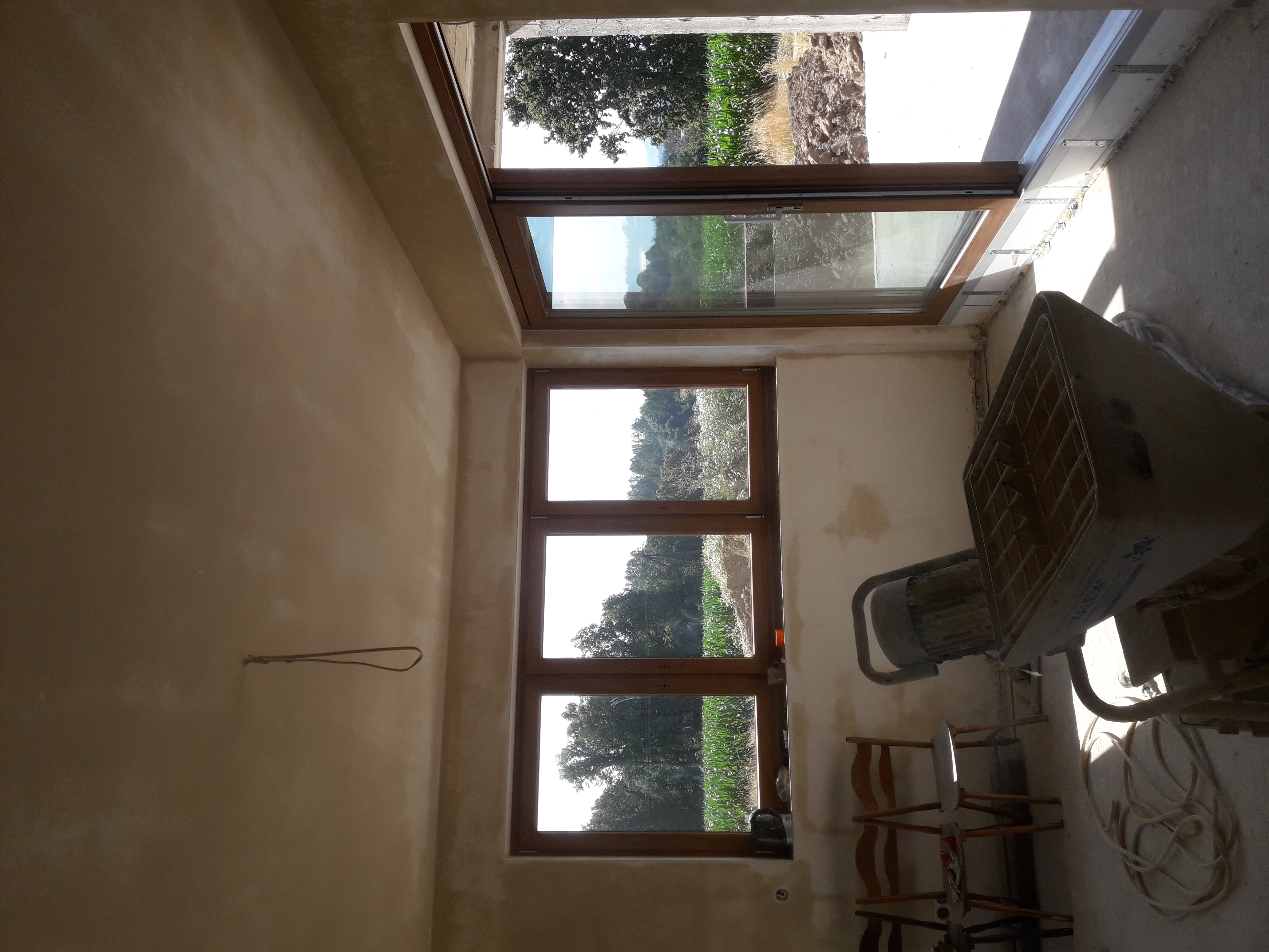 Wnętrze po nałożeniu jasnego tynku na ścianach, widoczne drewniane okna z widokiem na zewnątrz, mieszadło do tynku i krzesła w rogu pomieszczenia.