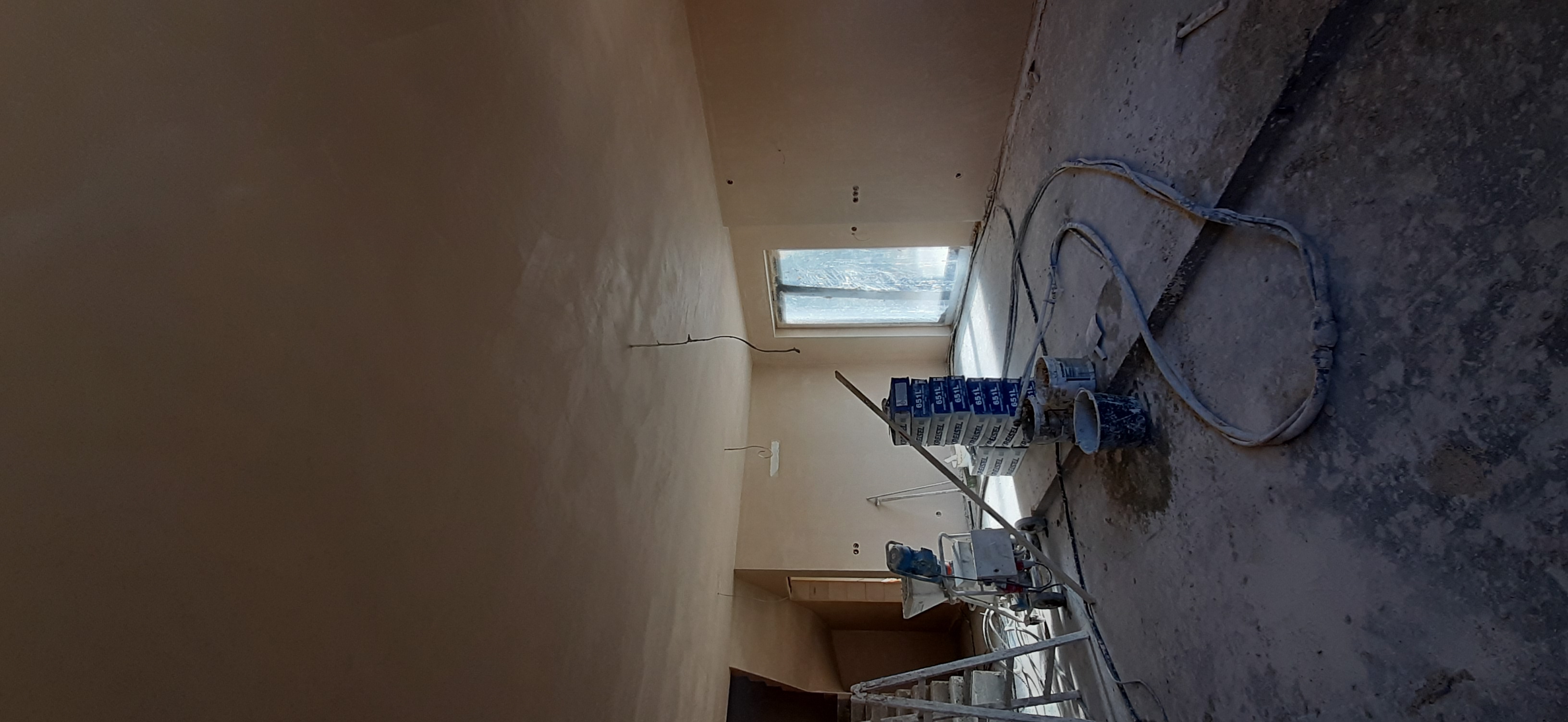 Świeżo otynkowana ściana w jasnym odcieniu, z widocznymi przewodami elektrycznymi, obok stos pudełek z zaprawą, węże i agregat tynkarski na zanieczyszczonej podłodze.