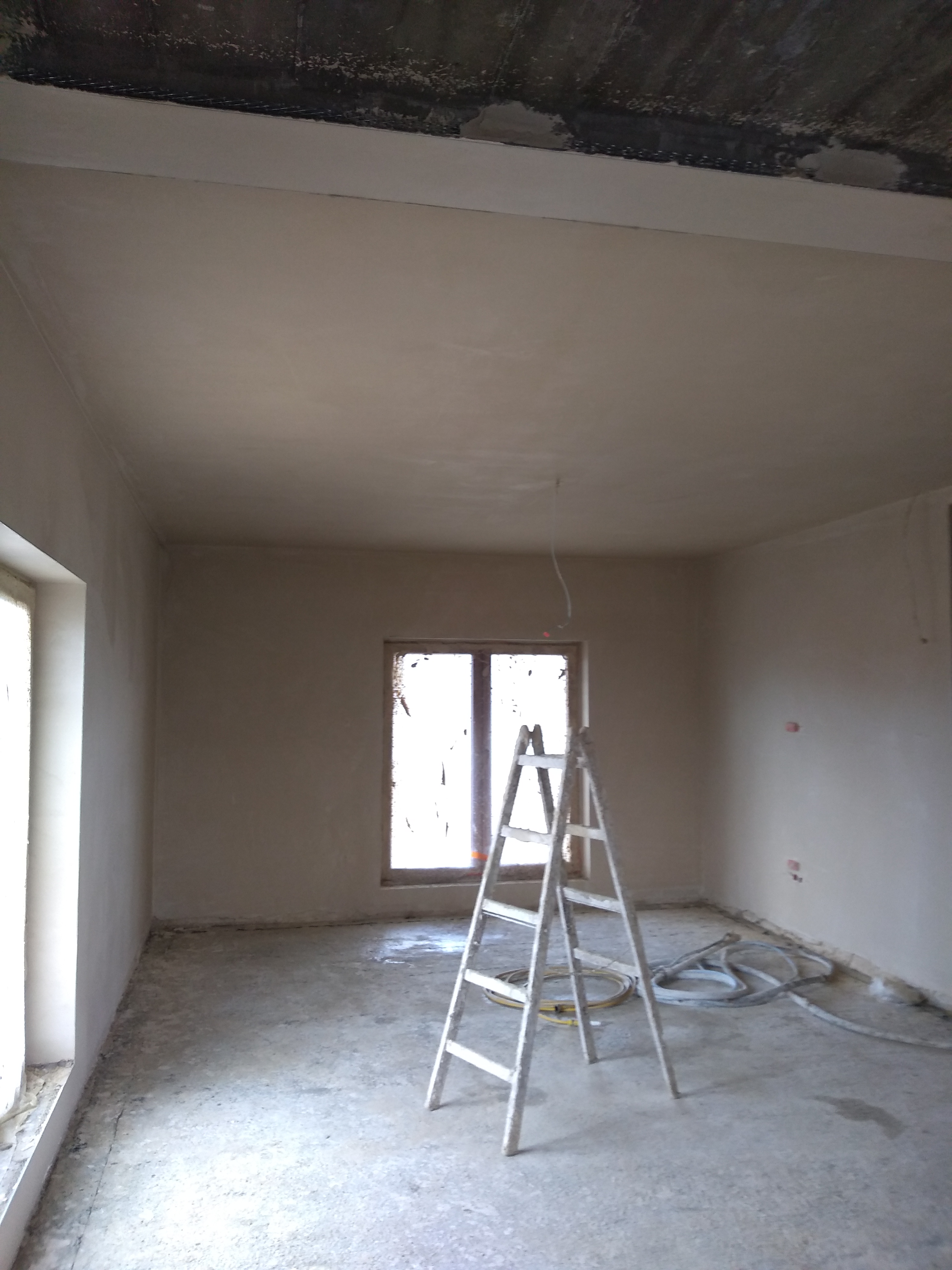 Wnętrze w trakcie tynkowania maszynowego, widoczna drabina, ściany i sufit pokryte świeżym tynkiem, fragment niezabezpieczonego sufitu, węże do tynkowania na podłodze.