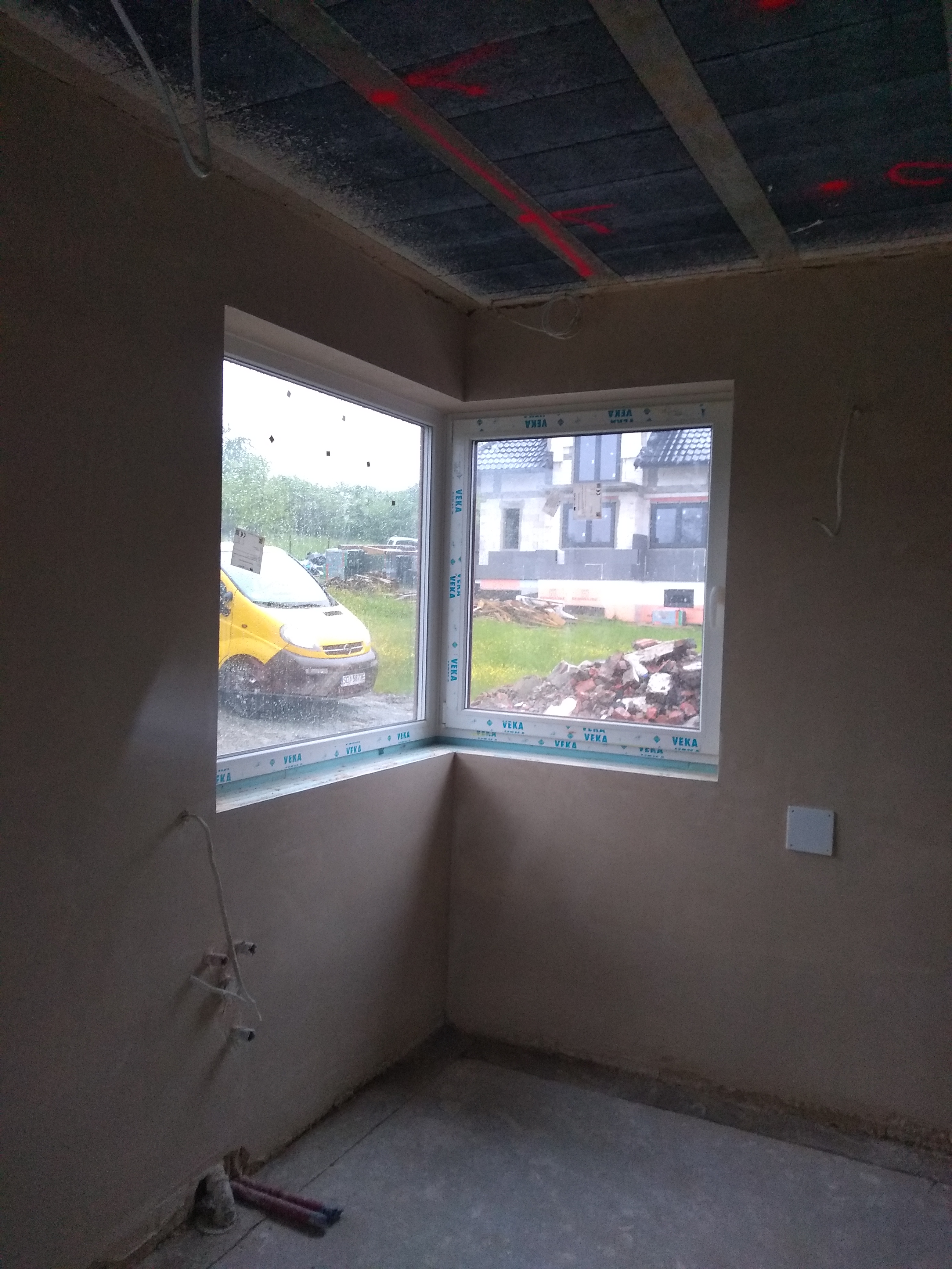 Świeżo otynkowane wnętrze pokoju z widocznymi przewodami elektrycznymi, oknami PCV marki VEKA i żółtym samochodem dostawczym za oknem w deszczowy dzień.