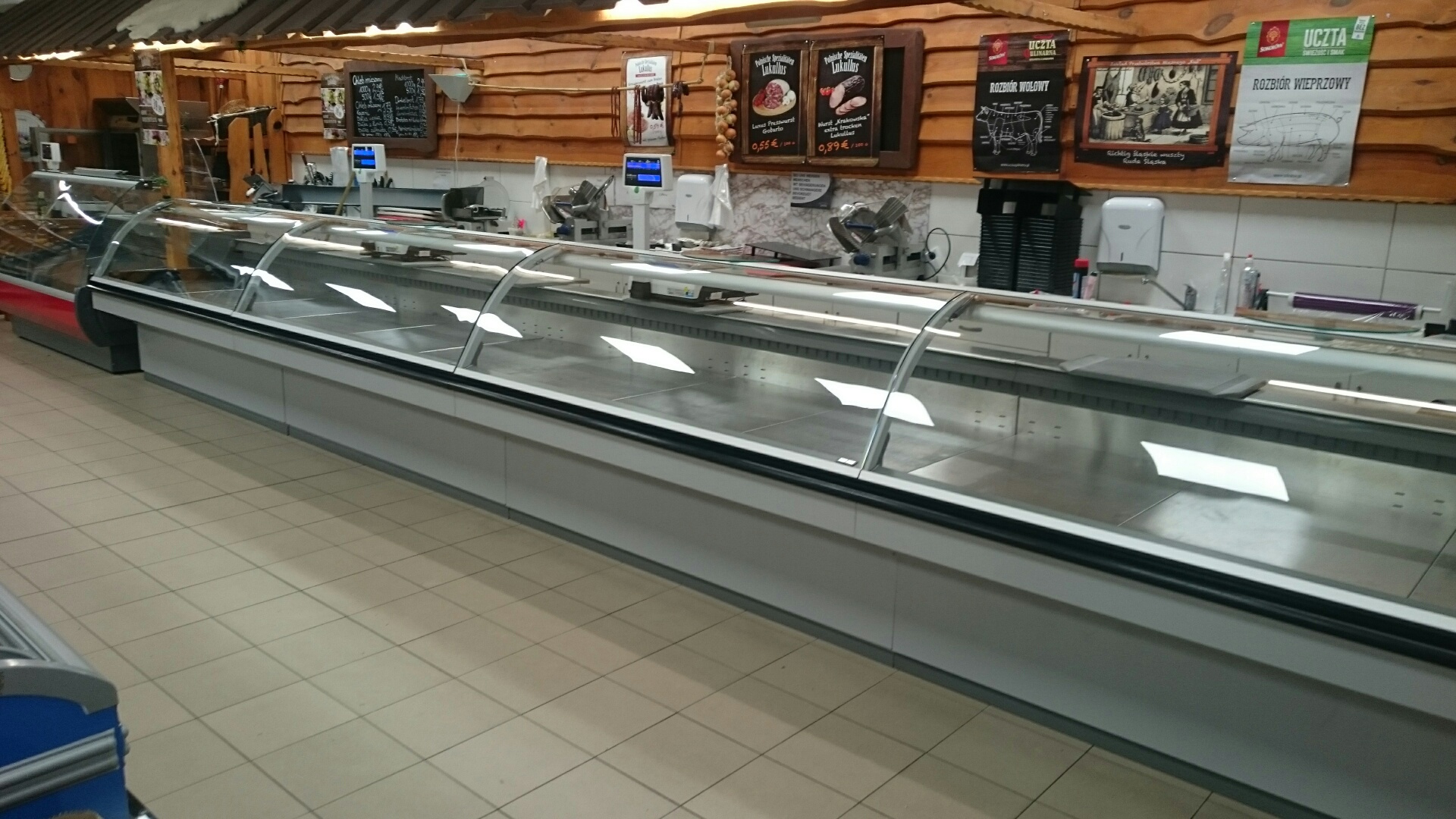 Długa, pusta chłodnicza lada ekspozycyjna z zakrzywioną szybą w sklepie mięsnym o wystroju rustykalnym, z drewnianymi panelami na ścianach i plakatami z rozbiorem wołowiny i wieprzowiny.