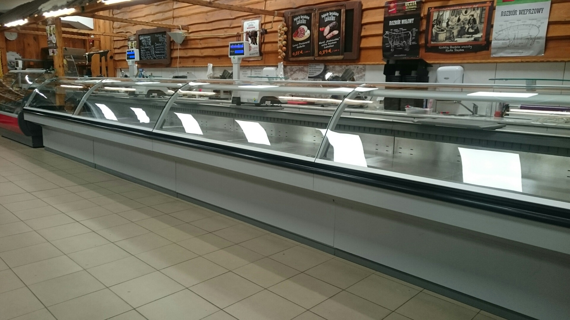 Pusta lada chłodnicza w sklepie mięsnym z drewnianym wykończeniem ścian i sufitu, widoczne tablice z cenami wędlin i schematami rozbioru mięsa.