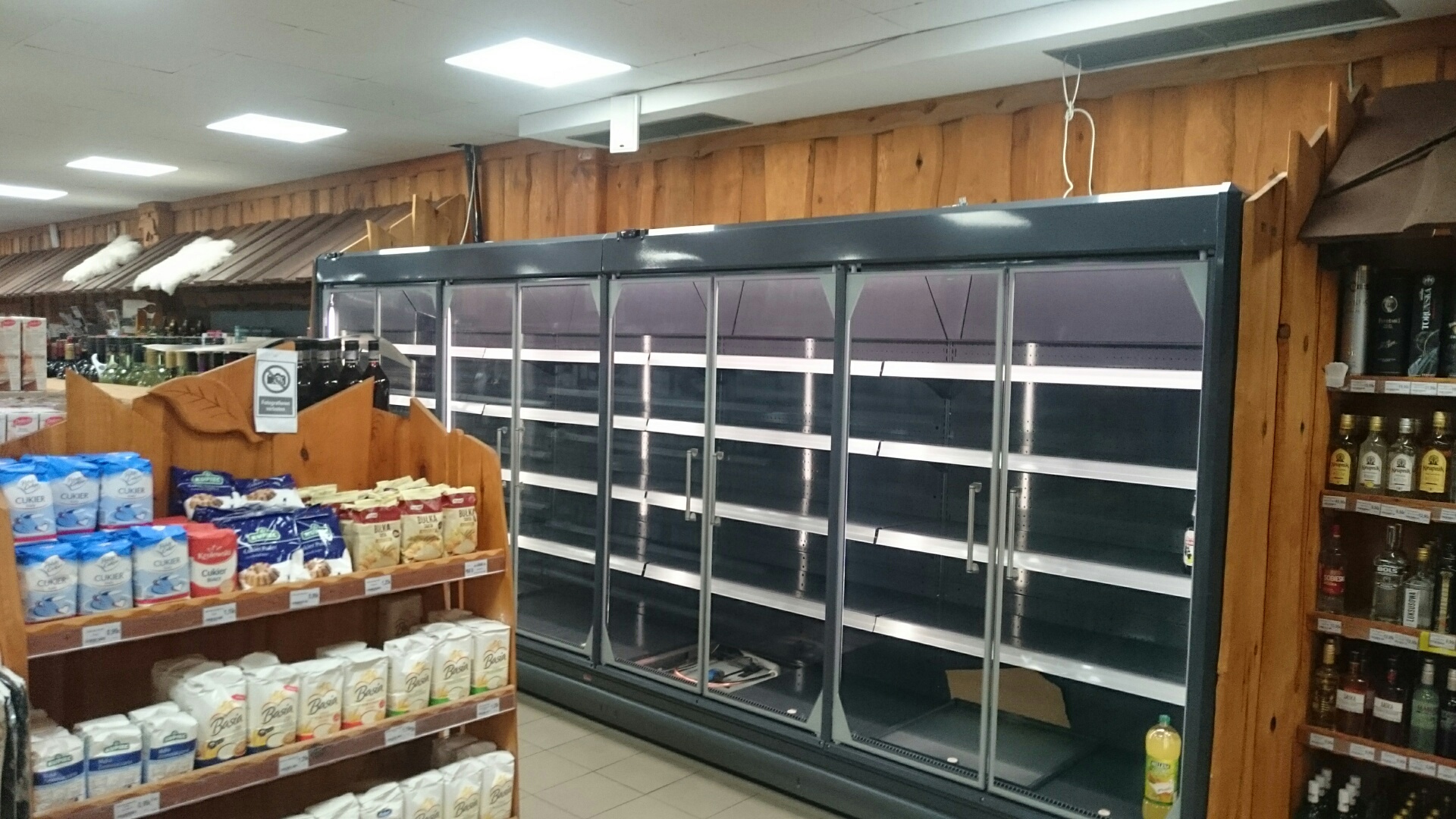 Półki w sklepie spożywczym: regał z mąką i cukrem po lewej, puste chłodziarki z przeszklonymi drzwiami w centrum, regał z alkoholem po prawej, drewniane wykończenie ścian i sufitu.