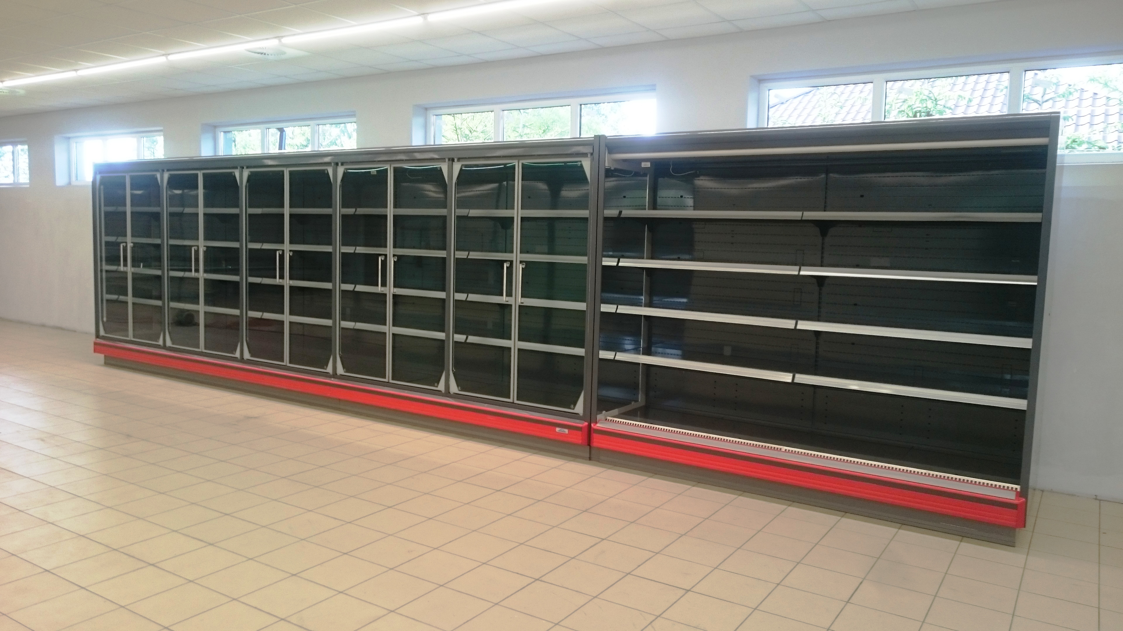 Pusta, szeroka chłodnicza lada sklepowa z przeszklonymi drzwiami i metalowymi półkami, stojąca na kafelkowej podłodze w jasnym pomieszczeniu z oknami.