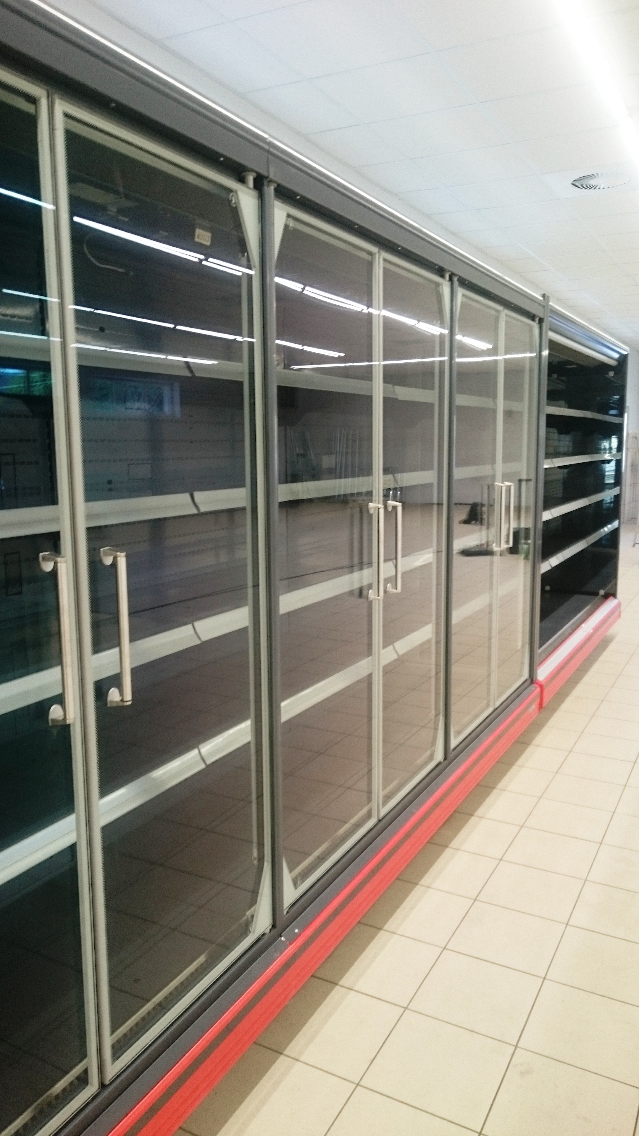 Długi, pusty regał chłodniczy z przesuwnymi szklanymi drzwiami i czerwonym akcentem u dołu, oświetlony jarzeniówkami, ustawiony na płytkach podłogowych w supermarkecie.