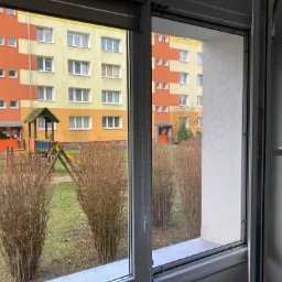 Widok z okna z zamontowaną moskitierą na tle bloku mieszkalnego z placem zabaw.