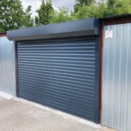 Szara, metalowa brama garażowa rolowana, zamontowana pomiędzy dwoma blaszakami, widok z zewnątrz.