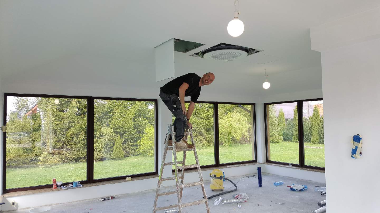 Mężczyzna na drabinie montuje klimatyzację sufitową w jasnym, przestronnym pomieszczeniu z dużymi oknami; widoczne narzędzia i niedokończone prace.