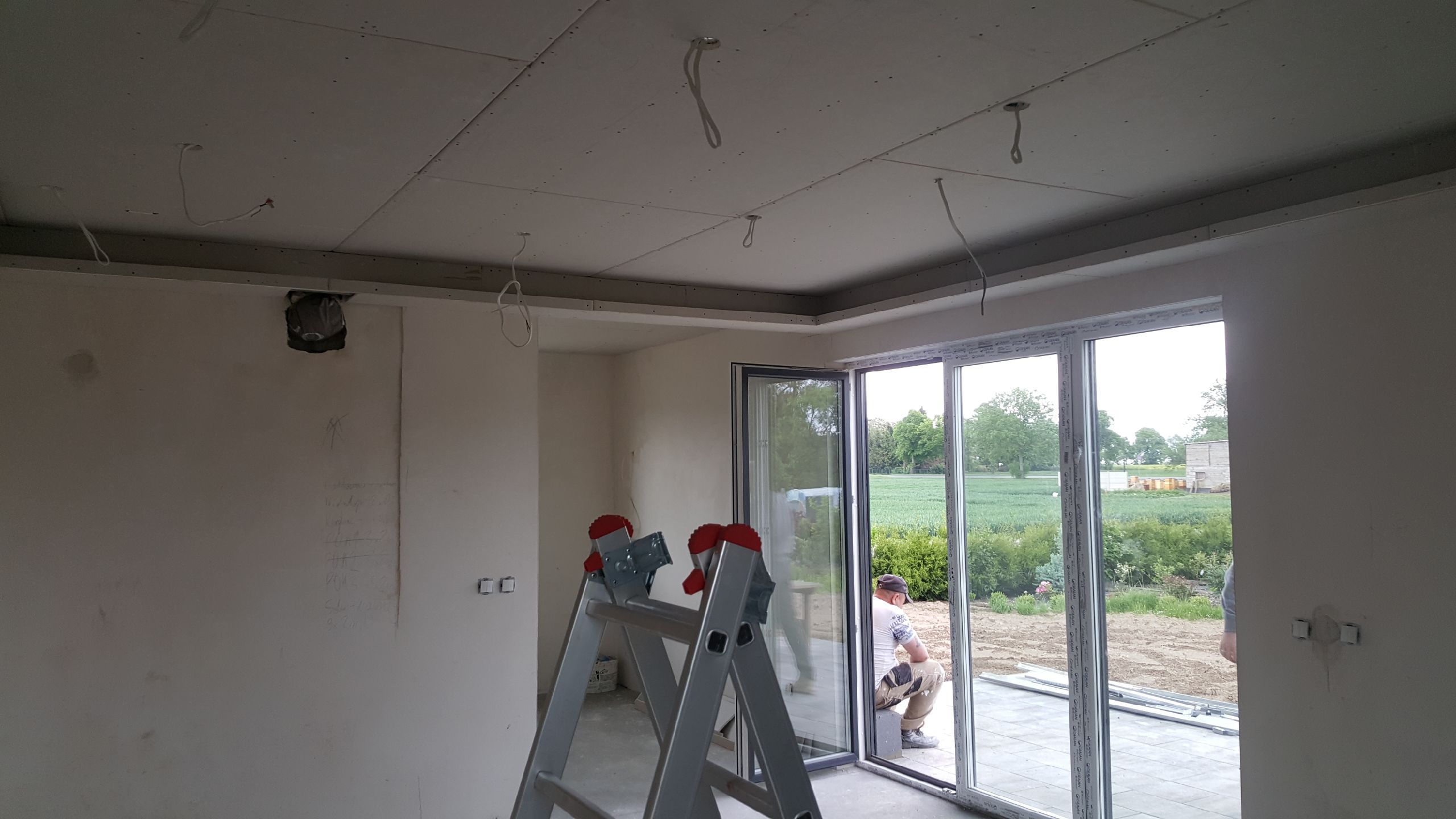 Wnętrze w trakcie prac wykończeniowych: widoczna surowa ściana z notatkami, częściowo zamontowana płyta karton gips na suficie z wystającymi kablami, aluminiowa drabina i pracownik odpoczywający...