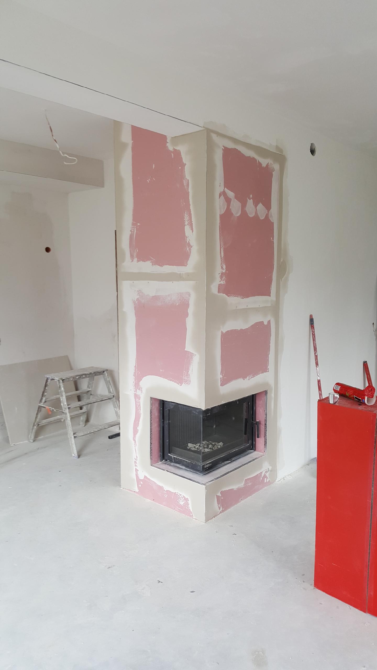 Wnęka kominkowa w trakcie obudowy płytami kartonowo-gipsowymi, widoczna metalowa kaseta kominka, surowe ściany i podłoga, drabinka malarska w tle.