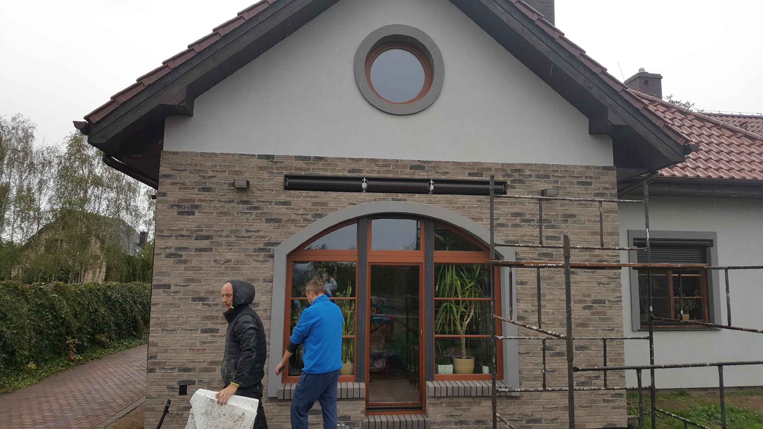 Elewacja domu z cegły klinkierowej w trakcie wykończenia, widoczne rusztowanie, okrągłe okno na poddaszu i pracownicy.