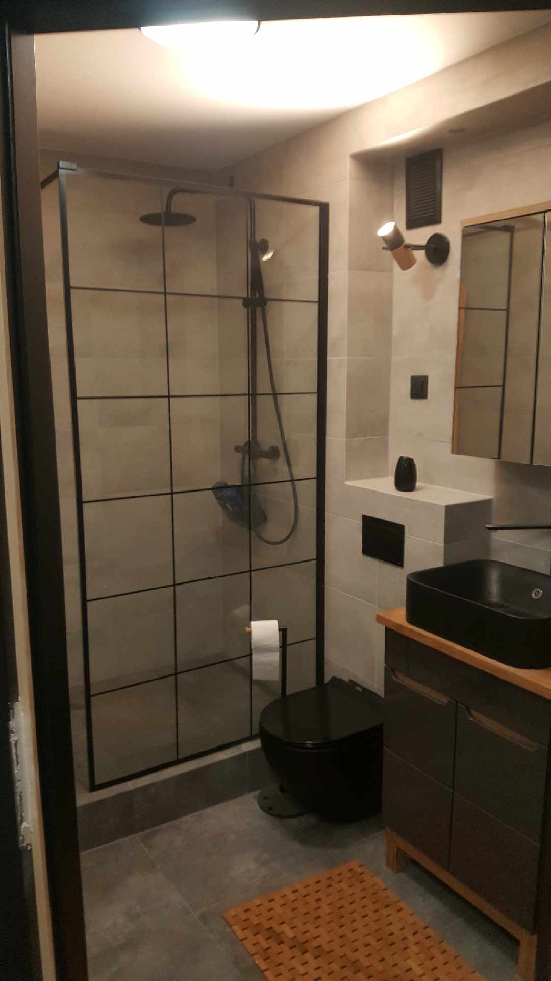 Nowoczesna łazienka z kabiną prysznicową w czarnej ramie, czarną umywalką nablatową i toaletą, szare płytki i drewniane akcenty.