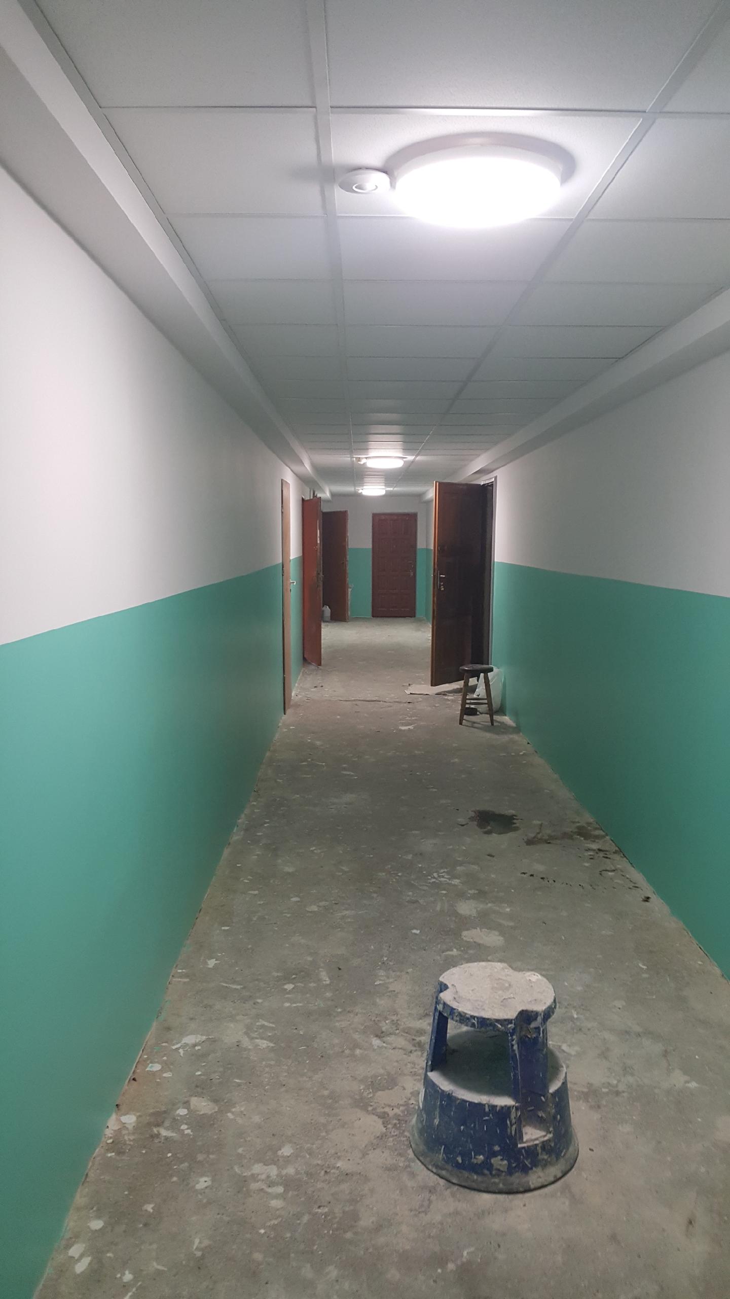 Korytarz w trakcie malowania, ściany pomalowane do połowy na zielono, powyżej białe, widoczne otwarte drzwi i stołek malarski.