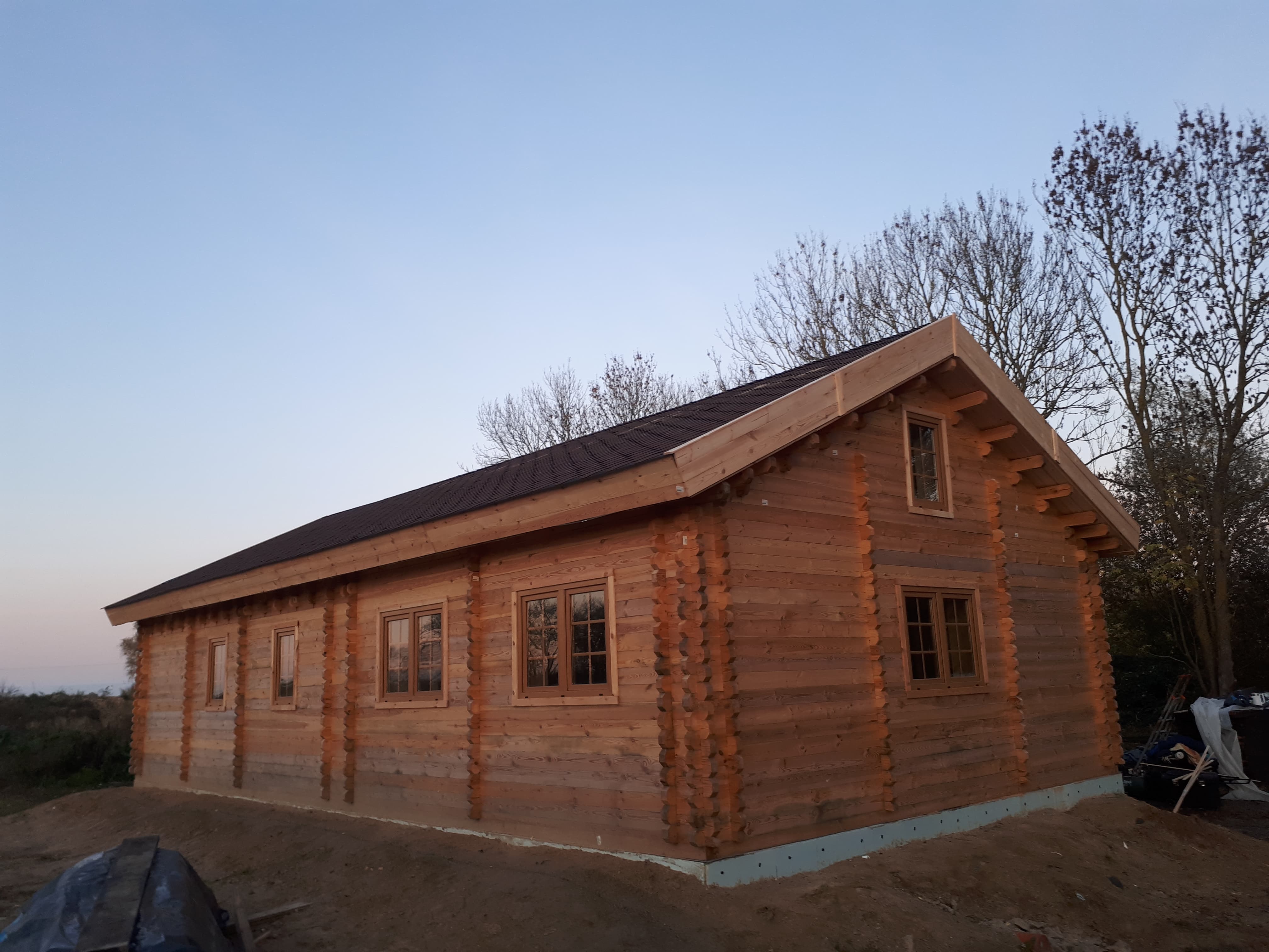 Nowoczesna altana z bali drewnianych z brązowym dachem, widok z zewnątrz, na etapie wykończenia terenu wokół.