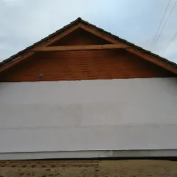 Surowy stan elewacji bocznej domu murowanego z drewnianą podbitką dachową i widoczną konstrukcją więźby dachowej, fragment sąsiedniej posesji w tle.