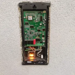 Otwarta obudowa urządzenia elektronicznego z widoczną płytką drukowaną, przewodami i elementami montażowymi, umieszczona w ścianie o szorstkiej fakturze.