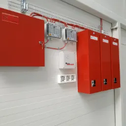 Czerwone szafy sterownicze i skrzynki elektryczne zamontowane na białej ścianie z poziomych paneli, instalacja przewodów w korytkach kablowych pod sufitem.