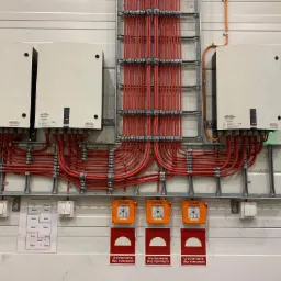 Rozbudowana instalacja elektryczna z licznymi przewodami w metalowych korytkach, panele sterujące urządzeniami oddymiającymi, schemat instalacji i przełączniki na ścianie z paneli.