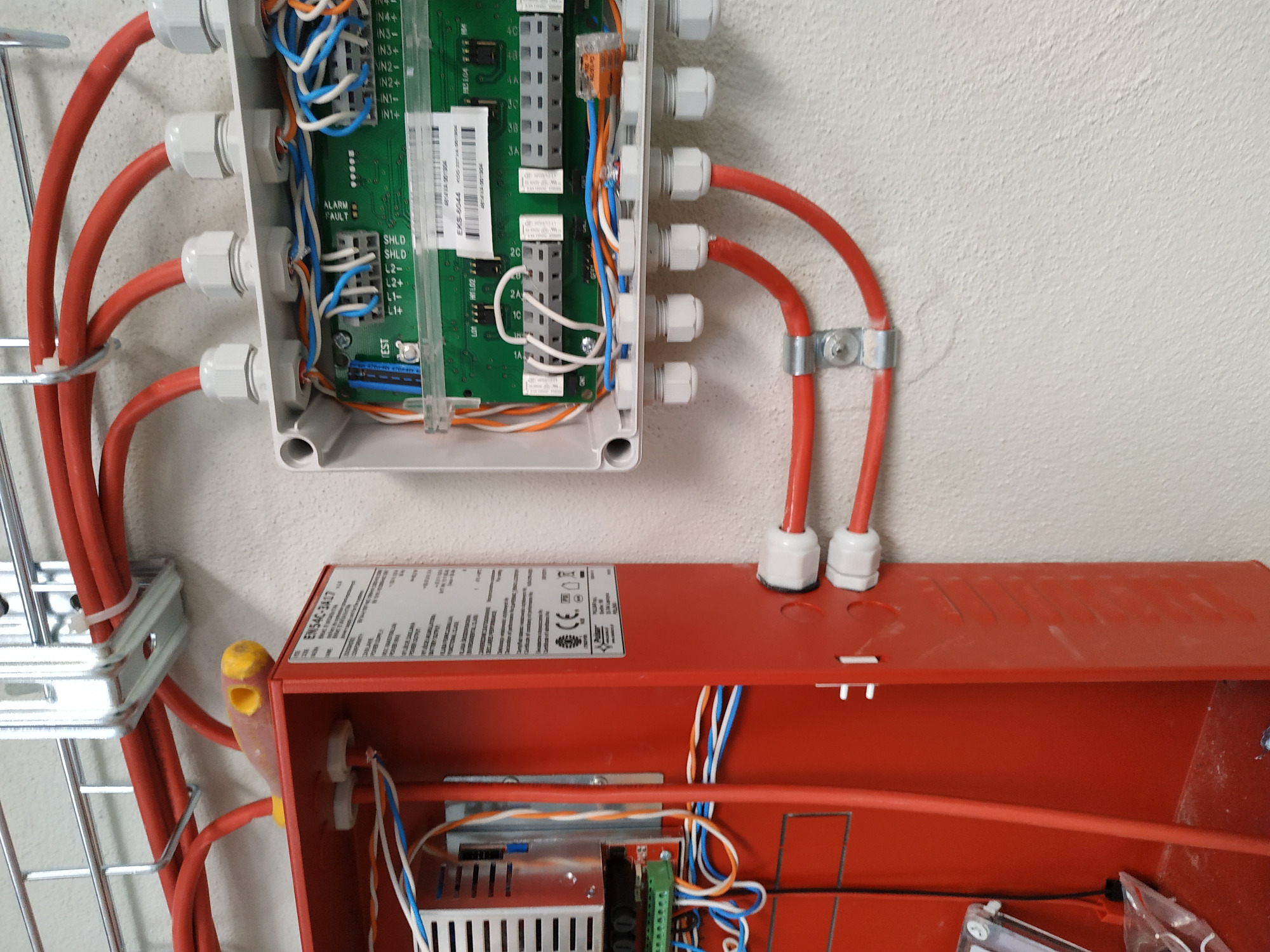 Otwarta skrzynka elektryczna z widoczną płytką drukowaną i podłączonymi przewodami w kolorze czerwonym, zamontowana na ścianie, poniżej czerwona obudowa z elektroniką i kablami.