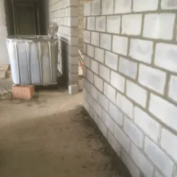 Wnętrze w trakcie budowy z widocznymi ścianami z białych bloczków betonowych, zbiornik na wodę i fragment pracującego murarza w pomarańczowej kamizelce odblaskowej.