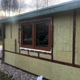 Ocieplenie ściany zewnętrznej budynku wełną mineralną, widoczne brązowe ramy konstrukcyjne i okno z pianką uszczelniającą, drabina oparta o ścianę, w tle drzewa.