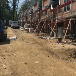 Wczesny etap budowy szeregu domów z czerwonej cegły, z widocznymi rusztowaniami, materiałami budowlanymi i pojazdami na placu budowy w słoneczny dzień.