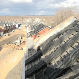 Panorama z budowy szeregu domów w stanie surowym otwartym, widoczne przygotowanie dachu z łatami i membraną oraz ułożone dachówki, w tle krajobraz miejski.