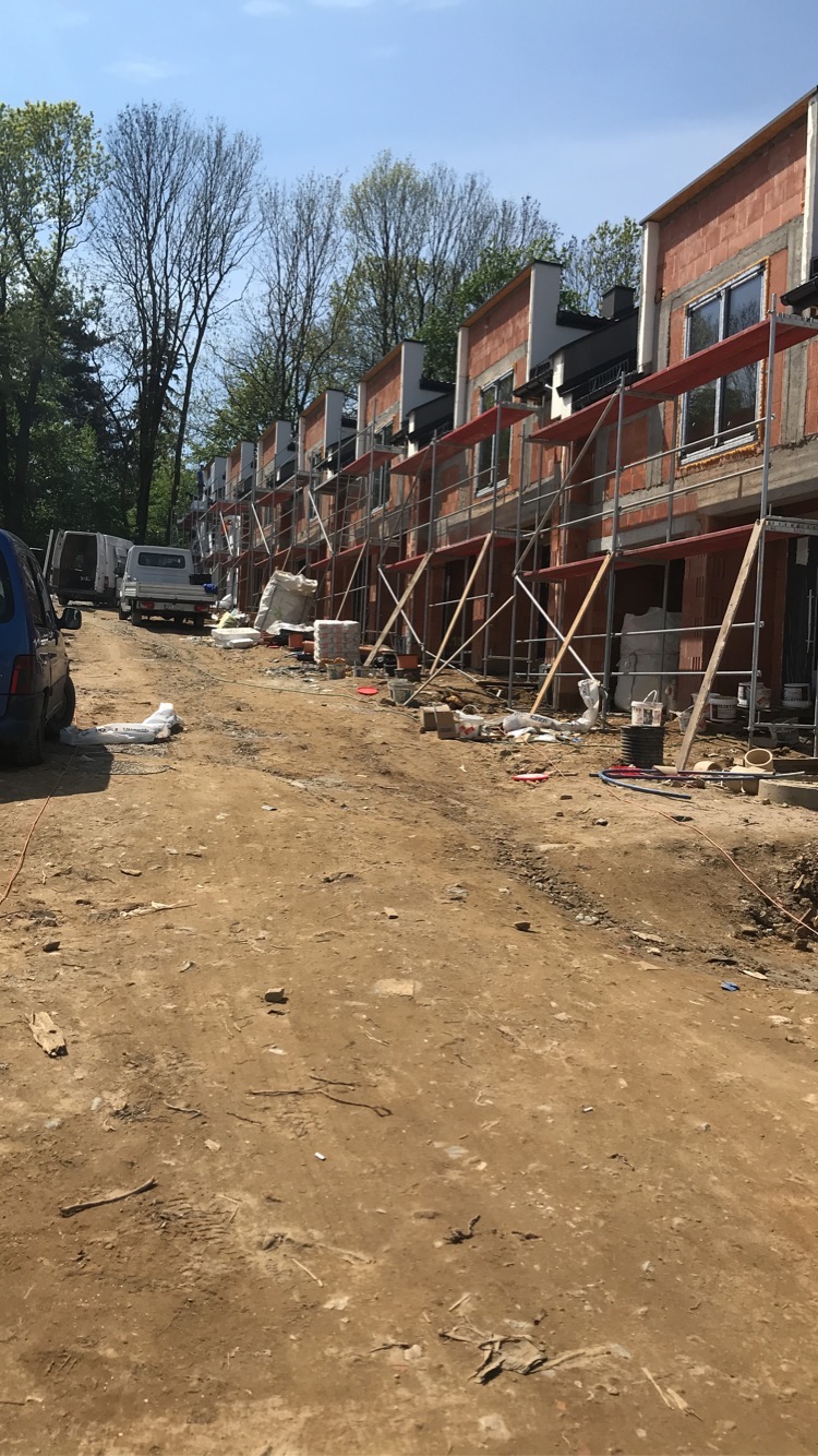 Wczesny etap budowy szeregu domów z czerwonej cegły, z widocznymi rusztowaniami, materiałami budowlanymi i pojazdami na placu budowy w słoneczny dzień.