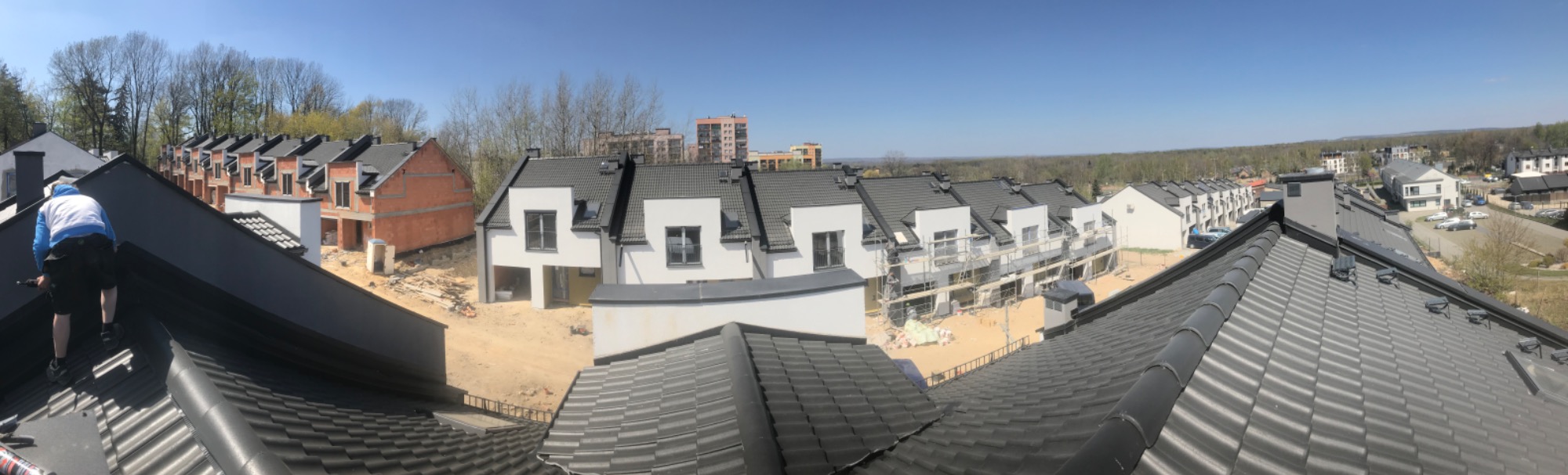 Pracownik w niebieskiej bluzie montuje ciemnoszare pokrycie na dachu szeregowca w trakcie budowy, widok z góry na osiedle domów w różnym stadium wykończenia.