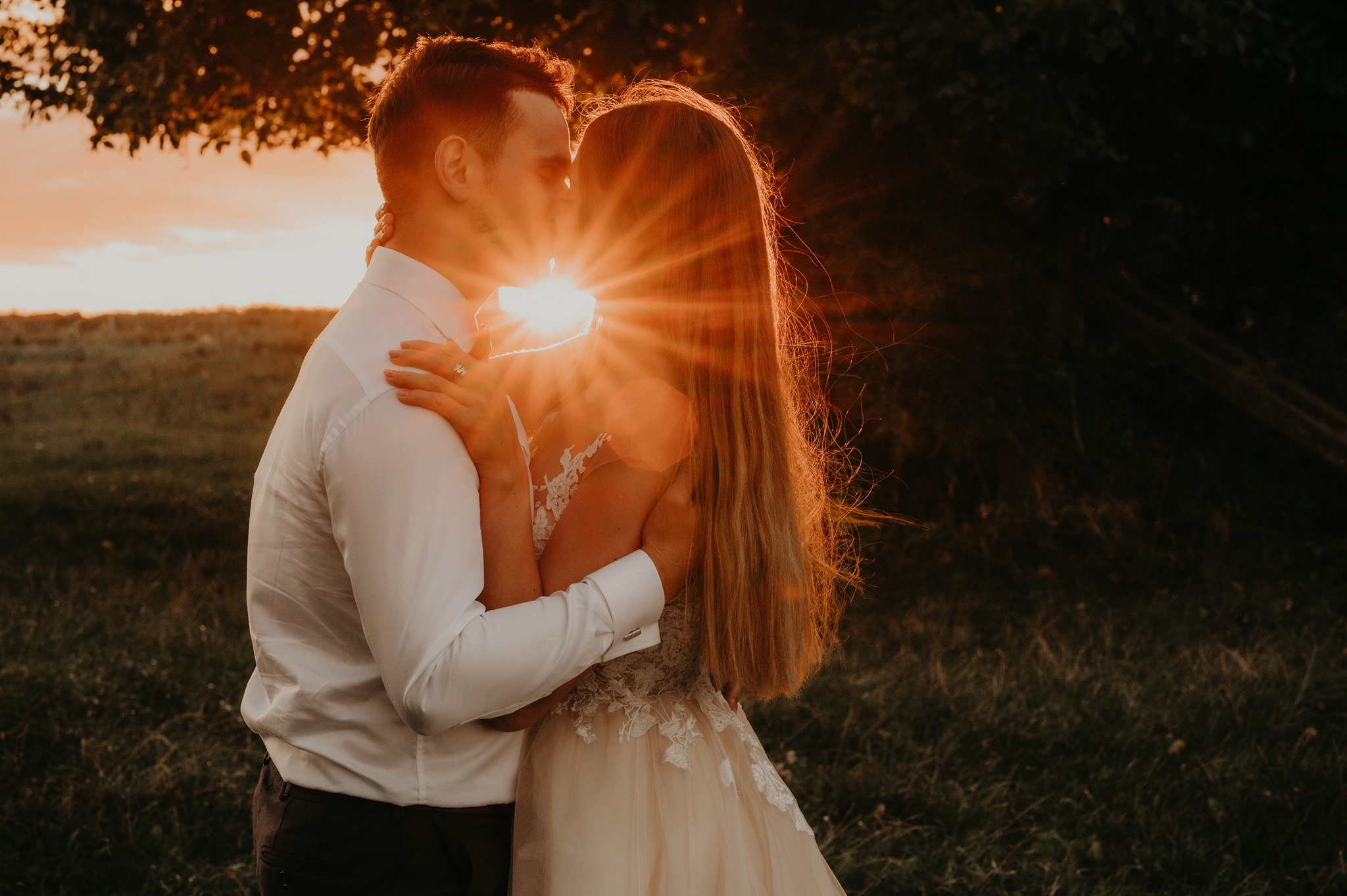 Para młoda w objęciach podczas pocałunku na tle zachodzącego słońca, pierścionek zaręczynowy widoczny na dłoni kobiety.