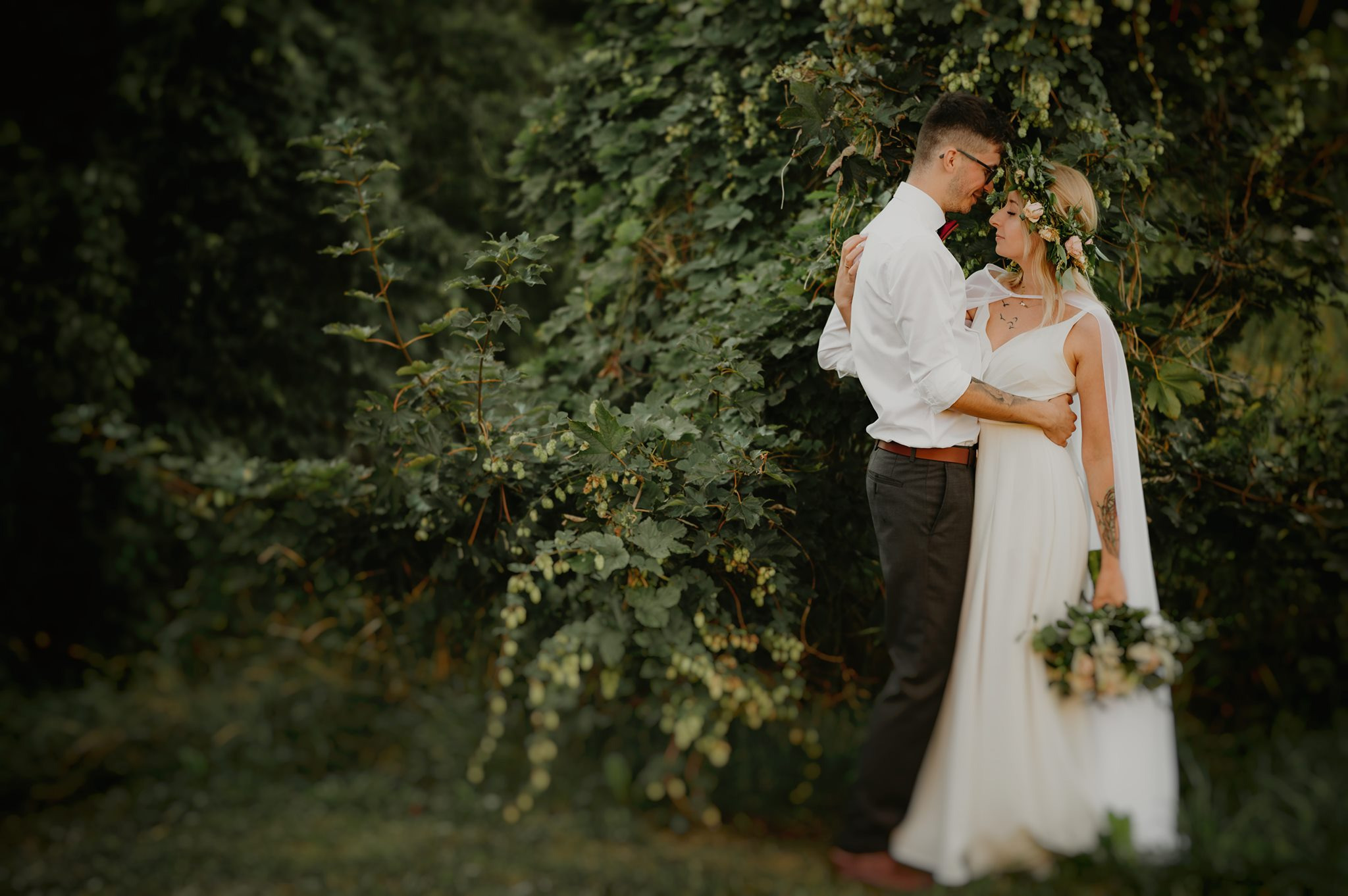 Para młoda w objęciach na tle gęstego, zielonego krzewu z owocami; panna młoda w białej sukni z wiankiem z kwiatów na głowie, pan młody w białej koszuli i bordowej muszce.
