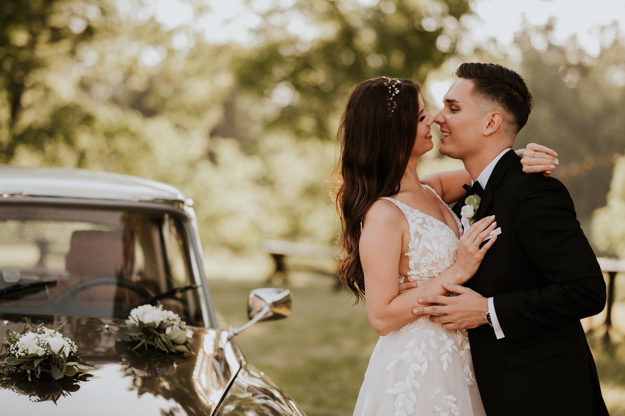 Uśmiechnięta para młoda w objęciach na tle klasycznego samochodu z bukietami kwiatów na masce, w tle zielone drzewa.