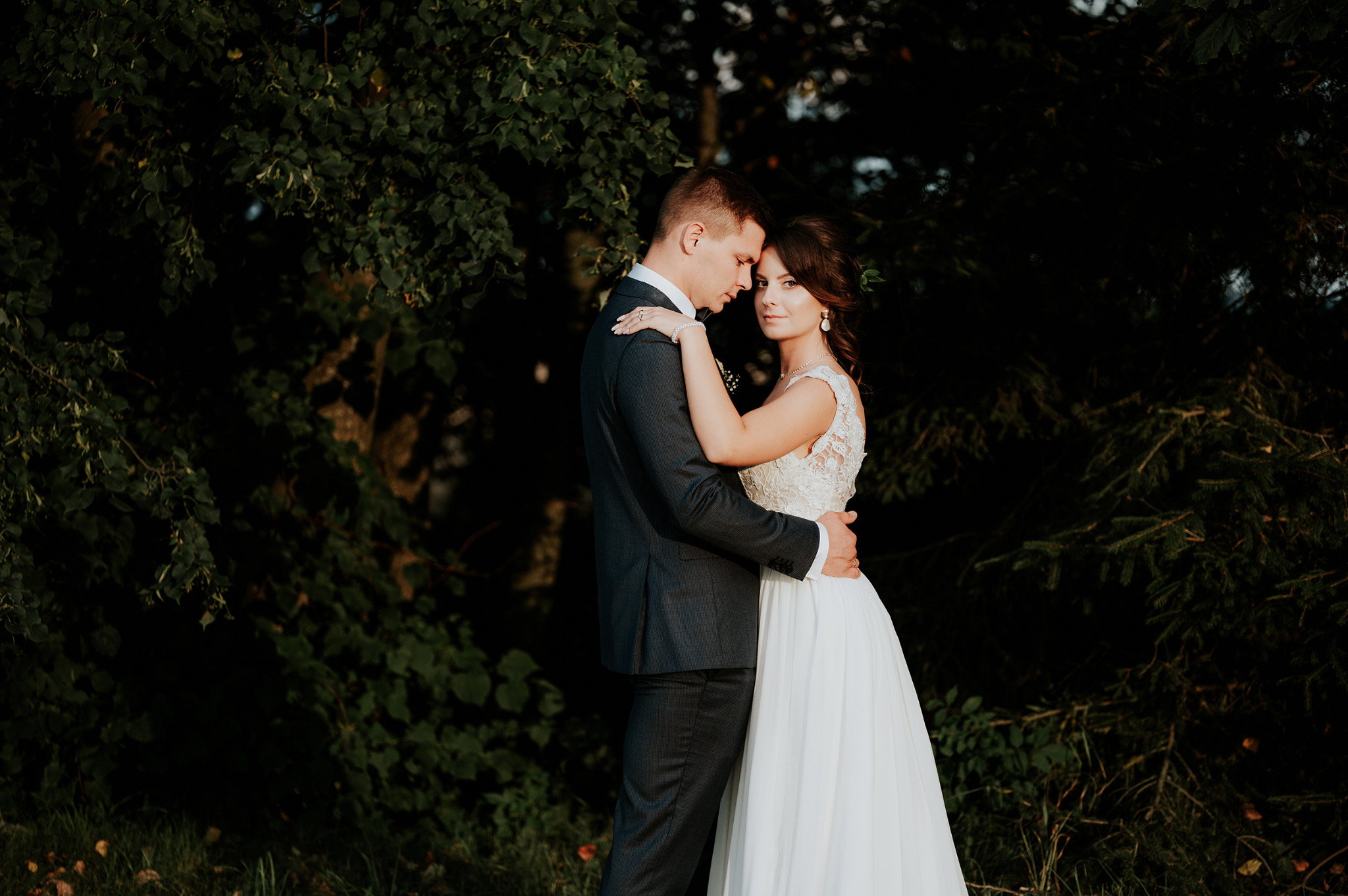 Para młoda w objęciach na tle ciemnej zieleni: panna młoda w białej sukni z koronkową górą i perłowymi kolczykami, pan młody w ciemnoszarym garniturze, delikatne światło podkreśla ich bliskość.