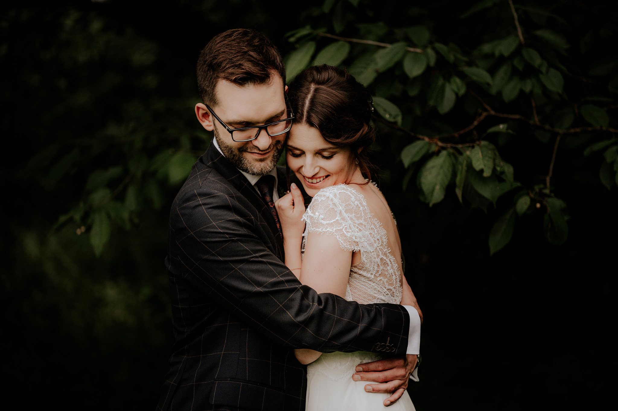 Uśmiechnięta para młoda w objęciach na tle ciemnej zieleni: on w garniturze w kratę i okularach, ona w koronkowej sukni ślubnej z odkrytymi plecami.