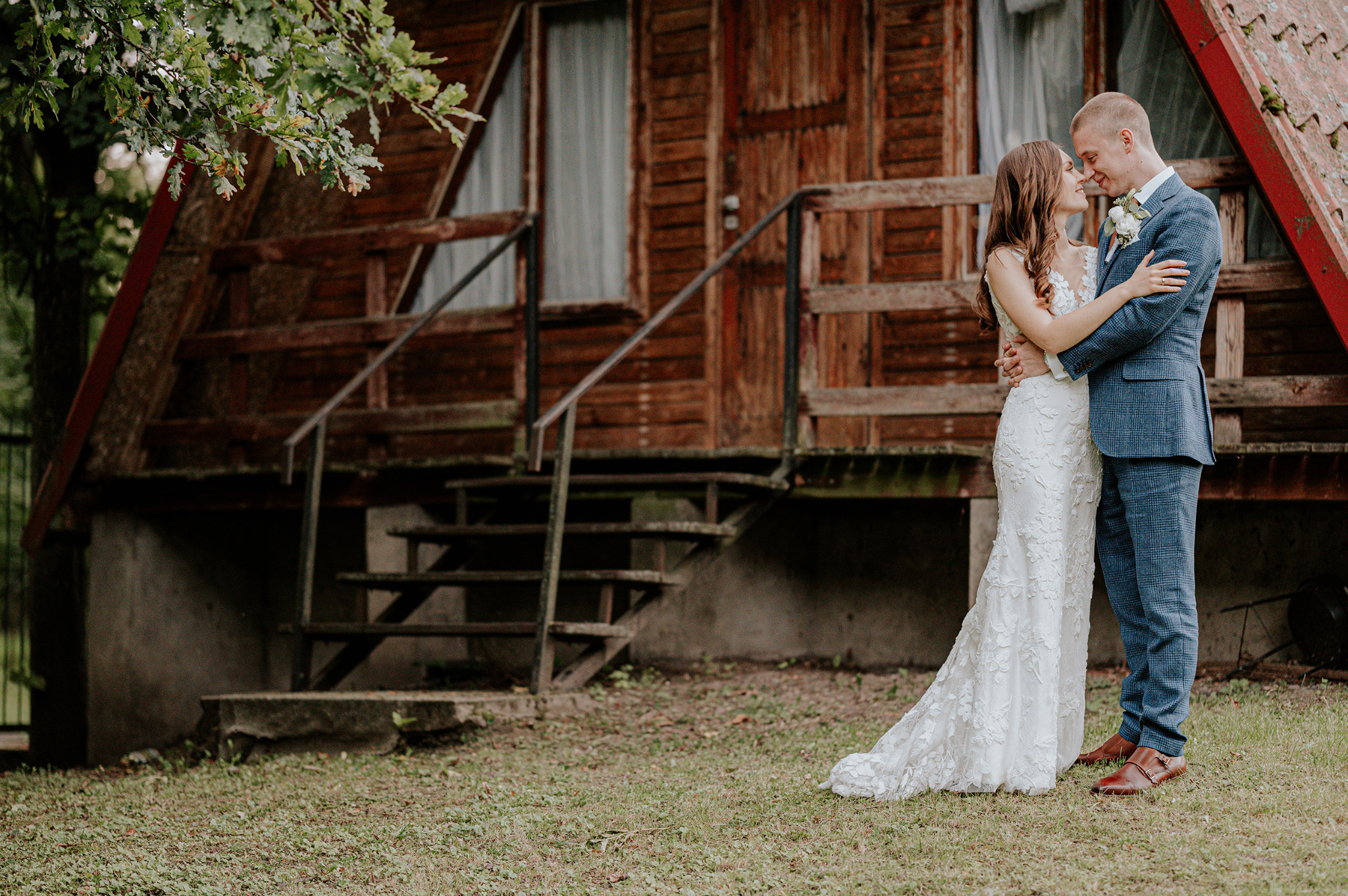Para młoda w objęciach przed drewnianym domkiem z czerwonym dachem, panna młoda w koronkowej sukni, pan młody w niebieskim garniturze w kratę.
