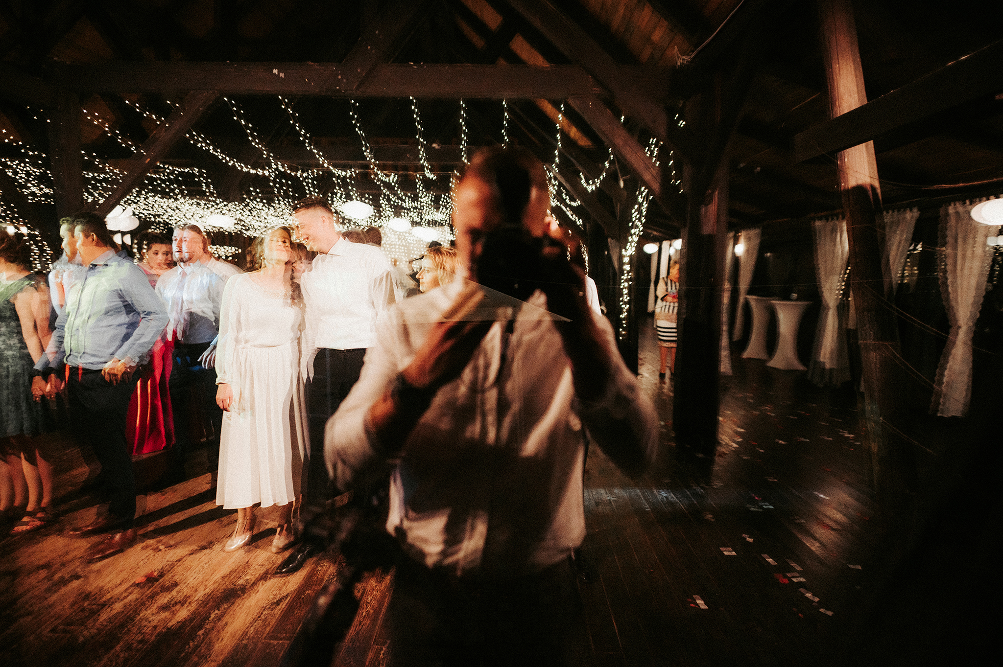 Goście w eleganckich strojach świętują na parkiecie pod drewnianą konstrukcją z girlandami światełek, fotograf rejestruje moment.