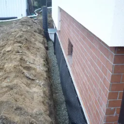 Wykop wzdłuż ściany budynku z cegły, zabezpieczony czarną folią i wypełniony żwirem. Widoczny drenaż pionowy i rura odprowadzająca wodę. Praca ziemna w toku.
