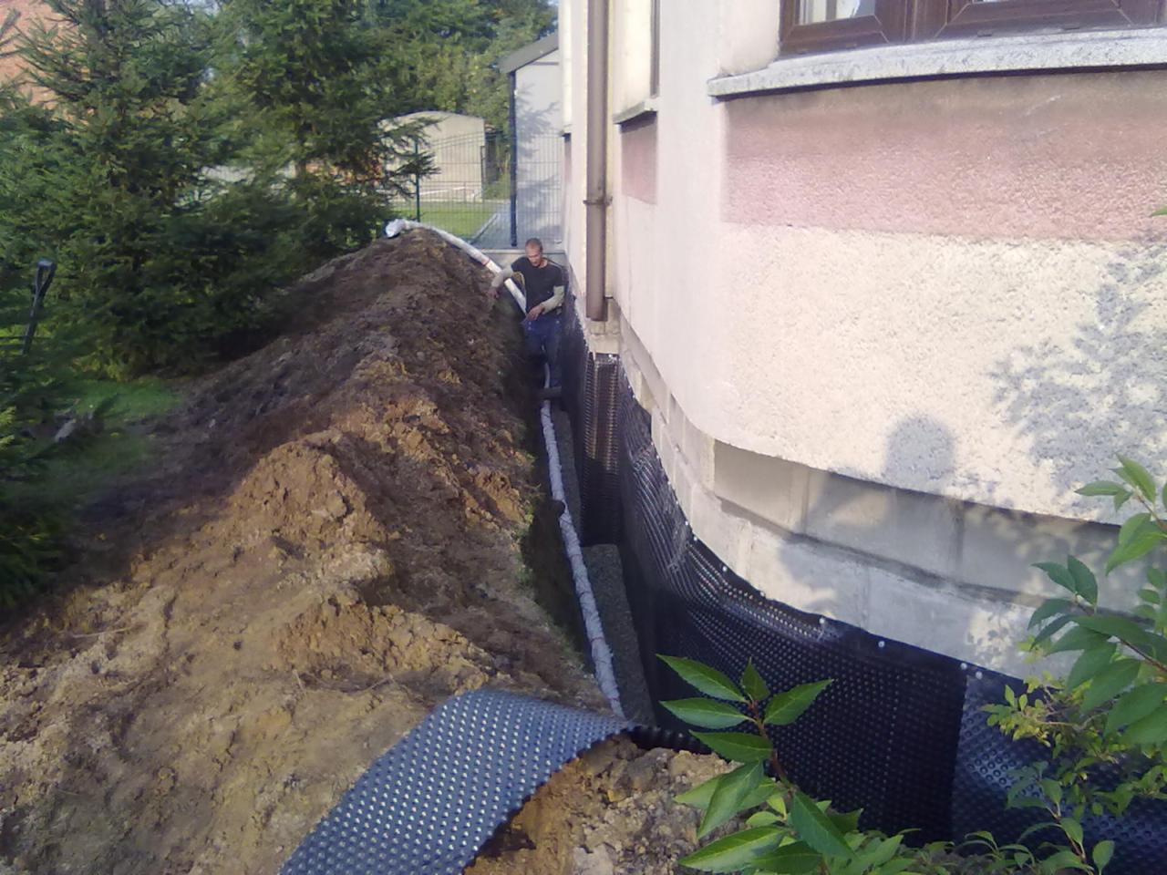 Odkopane fundamenty domu z izolacją pionową i drenażem. Pracownik w wykopie. Hałda ziemi. Drenaż odprowadza wodę rurą. Oświetlenie naturalne, dzień.