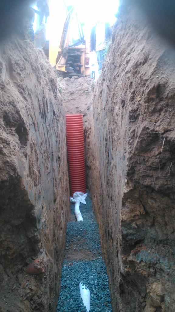 Wykop z rurą drenażową i podsypką żwirową. W tle widoczna koparka. Ściany wykopu z widocznymi warstwami ziemi. Ujęcie z dołu, podkreślające głębokość wykopu.