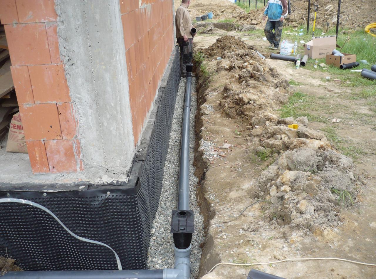 Drenaż fundamentów w trakcie montażu: rura drenażowa w wykopie, zabezpieczona folią kubełkową, widoczny fragment muru i osoby w tle.