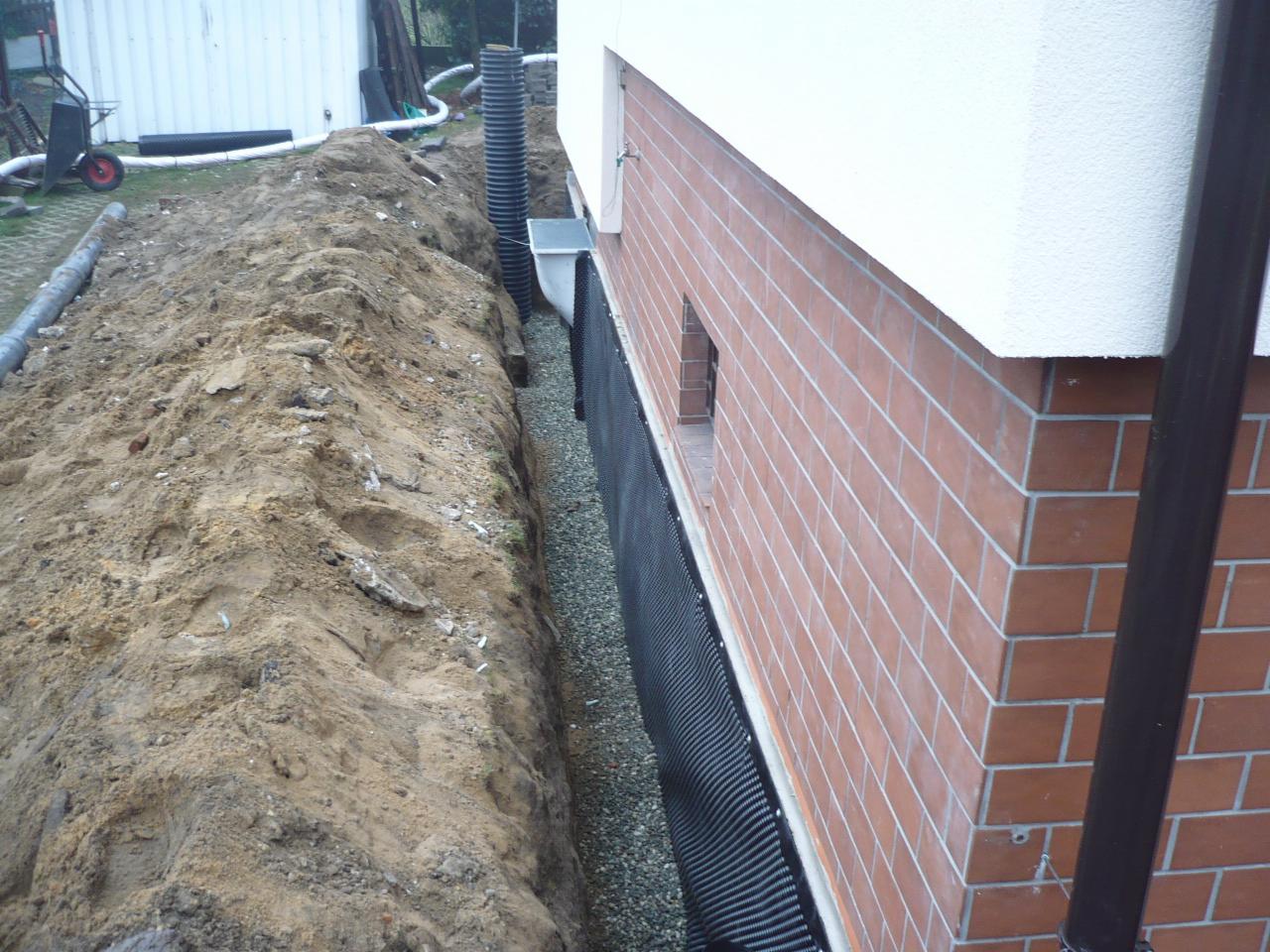 Wykop wzdłuż ściany budynku z cegły, zabezpieczony czarną folią i wypełniony żwirem. Widoczny drenaż pionowy i rura odprowadzająca wodę. Praca ziemna w toku.