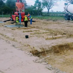 Wylewanie fundamentów na placu budowy z użyciem pompy do betonu i betonomieszarki; widoczne drewniane szalunki i pracownicy w odblaskowych kamizelkach.