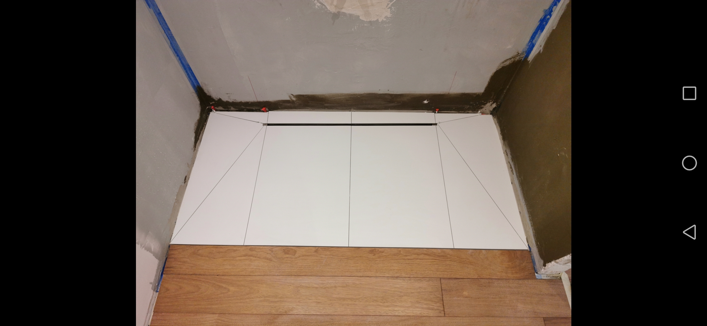 Układanie białych płytek na ścianie w trakcie prac, widoczne poziomowanie laserowe i plastikowe krzyżyki dystansowe, dolna część ściany wykończona drewnopodobnymi panelami podłogowymi.