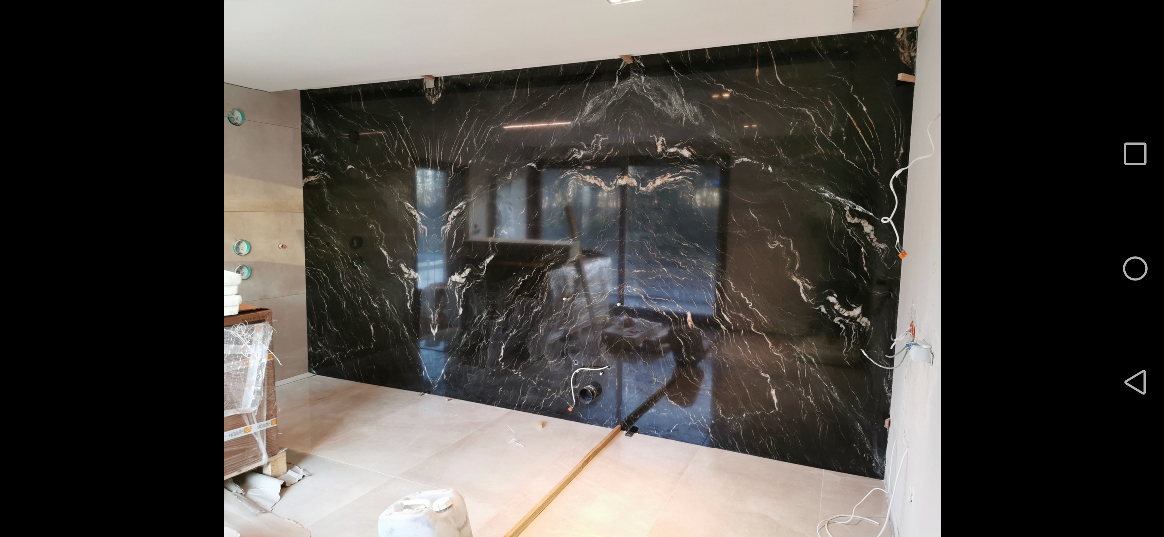Ściana w łazience wyłożona czarnym marmurem z białymi żyłami, widoczne przygotowane instalacje wodne i elektryczne, jasne płytki na podłodze, etap prac wykończeniowych.