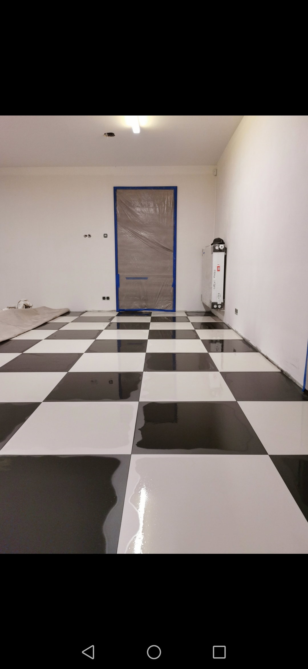 Nowo położona, błyszcząca podłoga w szachownicę, czarno-biała, z zabezpieczonymi folią i taśmą malarską drzwiami, widoczne niedoskonałości w postaci smug i nierówności na powierzchni.