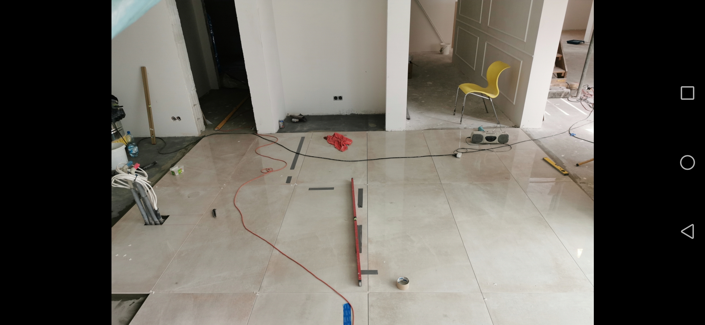 Układanie jasnych, błyszczących płytek podłogowych z użyciem poziomicy i krzyżyków dystansowych w przestronnym wnętrzu z widocznymi przewodami i otworem instalacyjnym.