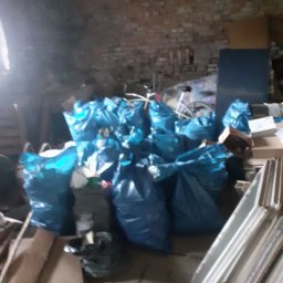 JUŻ w PORZĄDKU - Strych zagracony workami ze śmieciami, widoczne stare okna i ceglana ściana. Przygotowane do opróżniania, zbiórka odpadów w niebieskich workach.