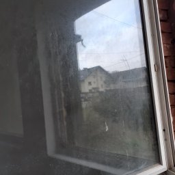 JUŻ w PORZĄDKU - Zabrudzone okno PCV z widokiem na budynki w Suchej Beskidzkiej. Widoczny kurz i zacieki na szybie. Okno uchylone, zarys ceglanej ściany w tle.