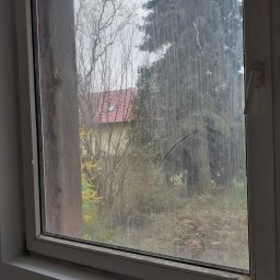 JUŻ w PORZĄDKU - Brudne okno z widokiem na zieleń i dom z czerwoną dachówką. Zabrudzenia na szybie utrudniają widoczność, rama okienna z widocznymi śladami użytkowania.