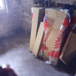 JUŻ w PORZĄDKU - Stare, surowe pomieszczenie z oknem, w którym składowane są płyty izolacyjne z folią ochronną. Widoczne ślady wilgoci na ścianach i podłodze.